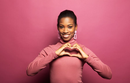 A young afro american woman laughs happily and shows a heart from her fingers. Portrait on a pink background.の写真素材