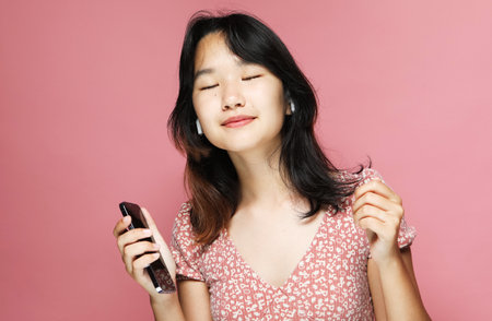 A young pretty Asian woman smiling and listening to music with yearphones, holding a mobile in her hand on a pink background,の写真素材