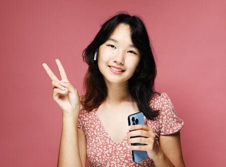 A young pretty Asian woman smiling and showing a peace sign while listening to music with...