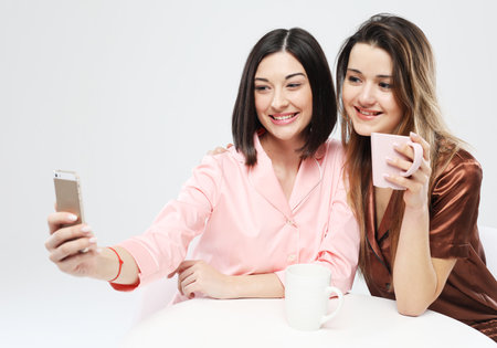 Two smiling cheerful female friends dressed in pajamas drink coffee and take selfies.