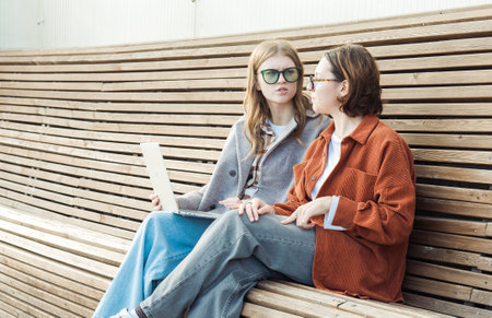 Two female student friends are sitting on a wooden bench outside with a laptop.の写真素材