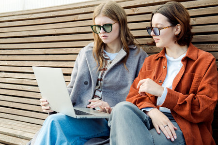Two female student friends are sitting on a wooden bench outside with a laptop and discussing their studies.の写真素材