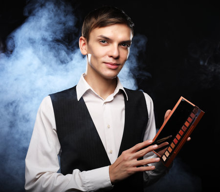 Professional makeup artist male posing in studio, holds a palette and brushes. Dark background with smoke. Fashion concept.の写真素材