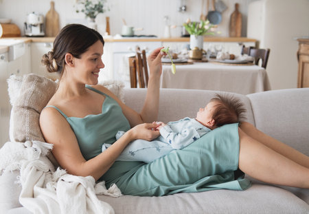 A young happy mother is sitting on the sofa and holding a baby in her arms.の写真素材