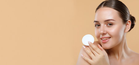 Woman using cotton pad over beige background. Happy smiling beautiful young female model cleaning skin by cotton pad.の写真素材