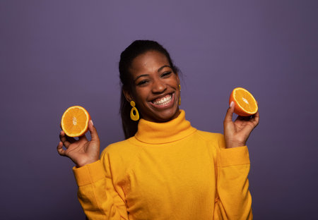 Charming dark-skinned young woman in a yellow sweater holds orange slices over purple background.の写真素材
