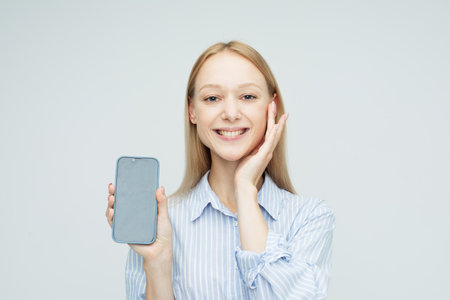 Portrait of a young smiling woman showing or presenting mobile phone over white background.の写真素材