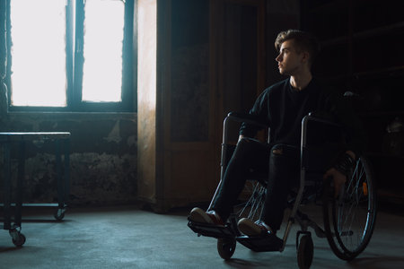 Young man sitting in a wheelchair in a dark room near window.の写真素材