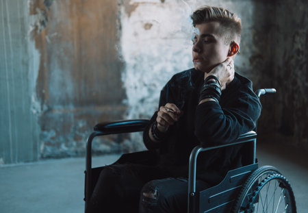 Depressed young man in a wheelchair smokes a cigarette, in dark room near window. Pain, fatigue.の写真素材