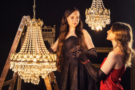 Two gorgeous female models in evening dresses and gloves in a beautiful room with a luxurious chandelier.の写真素材