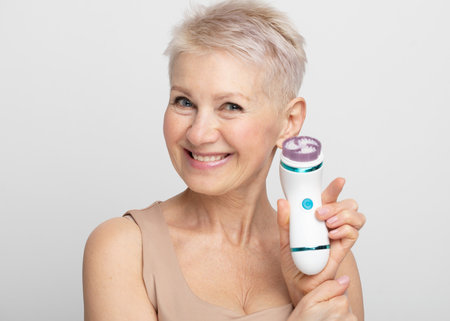 Beautiful 60s woman holding a massager in her hand over gray background. Spa technique skincare ads.の写真素材