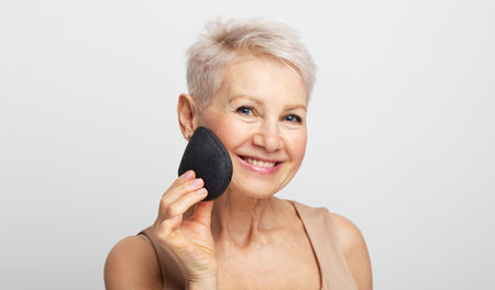 Charming old woman with short hair use sponge remove make up on light gray background, close upの写真素材