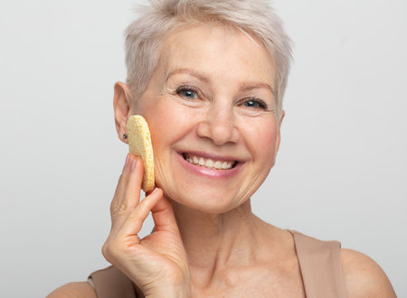 Charming old woman with short hair use sponge remove make up on light gray background, close upの写真素材