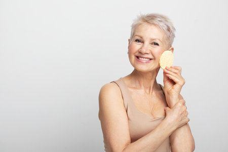 Charming old woman with short hair use sponge remove make up on light gray background, close upの写真素材