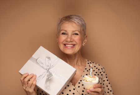 Cheerful elderly woman with short haircut holds a cupcake with a candle and present box, celebrates a birthday over beige background.の写真素材