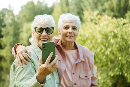 Two Caucasian senior women smiling to smartphone camera while taking selfie photos near lake in the...