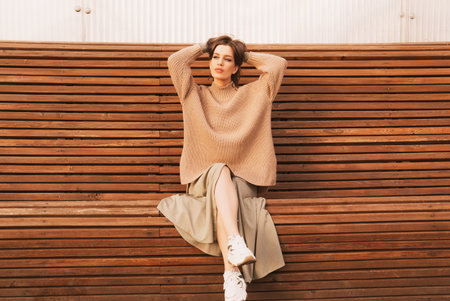 Young stylish woman model in an autumn look, in a sweater and skirt, sitting on a wooden bench. Fashion concept.の写真素材