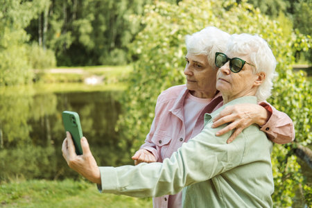 Two Caucasian senior women smiling to smartphone camera while taking selfie photos near lake in the park. Old ladies posing to phone.の写真素材