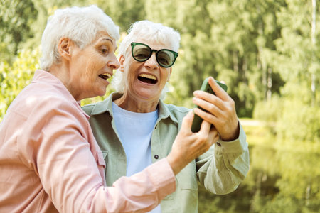 Two Caucasian senior women smiling to smartphone camera while taking selfie photos near lake in the park. Old ladies posing to phone.の写真素材