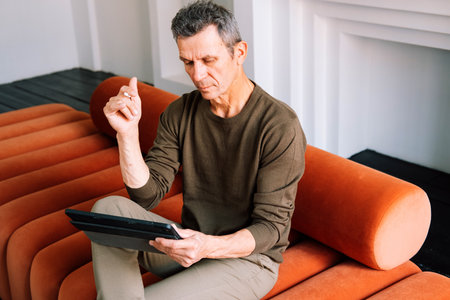 senior man in casual clothing using digital tablet while sitting on the sofa at homeの写真素材