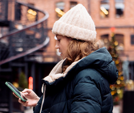 Adult woman in winter clothes and a hat holds a smartphone while typing a message in a winter city.の写真素材