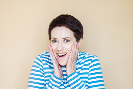 Shocked adult woman in striped shirt, looking at camera over beige background.