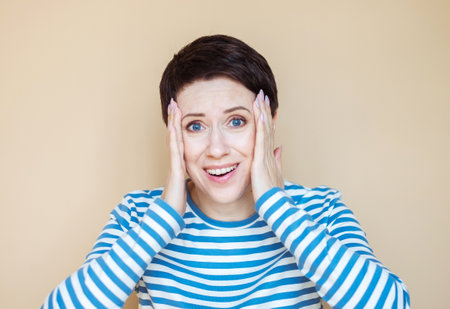 Shocked adult woman in striped shirt, looking at camera over beige background.の写真素材