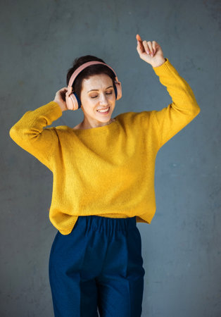 Woman in yellow sweater listening to music on headphones and dancing over grey background.の写真素材