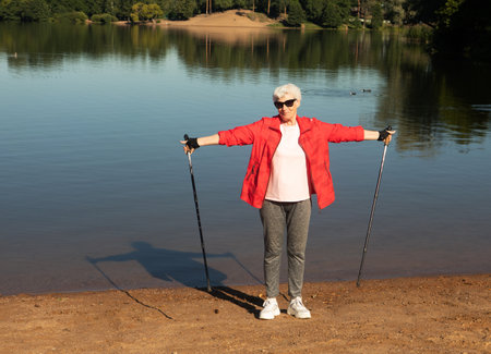 Grey-haired elderly woman walking with tracking sticks on the beach near lake. Summer time.の写真素材