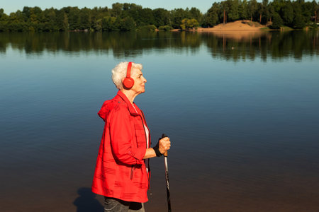 Elderly happy woman with gray hair and a short haircut in a red jacket walks with trekking poles and listens to music near lake.の写真素材