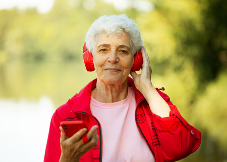 An elderly smiling woman with a short haircut dressed in red sportswear listens to music on headphones in the summer park.の写真素材