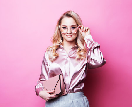 Beautiful young blond woman in a blouse and pants wearing glasses, holding handbag posing over pink background.の写真素材