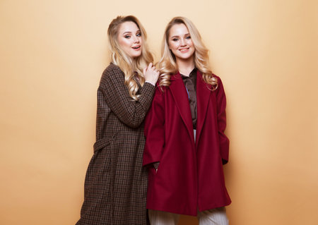 Positive portrait of two sisters, best friends posing indoor on beige background wearing winter stylish coat. Fashionable clothes.の写真素材