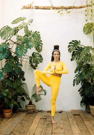 Young woman wearing yellow sportswear with headphones does yoga against the background of green plants.の写真素材