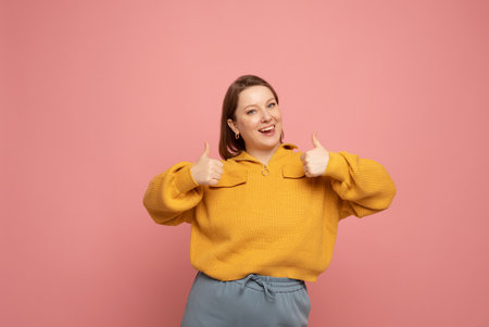 Portrait of cheerful woman in yellow sweater showing thumbs up gesture, isolated on pink background.