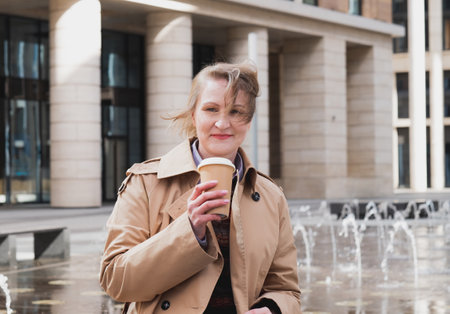 Beautiful mature blond woman in beige coat wit coffee to go in modern city.の写真素材