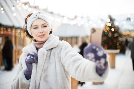Charming young woman in winter clothes at the Christmas market uses a smartphoneの写真素材