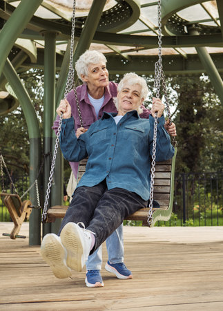 A cheerful happy old sisters is having fun in a summer park swinging on a swing.の写真素材