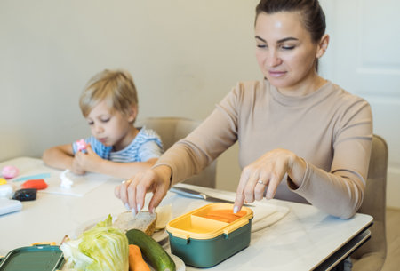 Young woman packs a lunchbox for her six-year-old son to take to school. Mother and boy in the kitchen in the morning.の写真素材