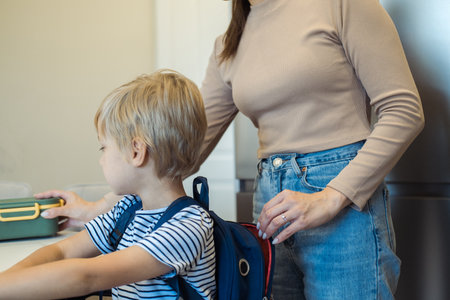 A woman puts a lunchbox into her seven-year-old son's backpack. Hands close up.の写真素材