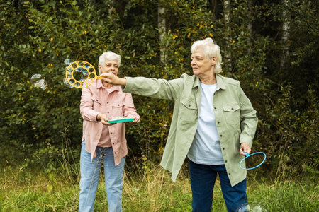 Two elderly sisters blowing soap bubbles near lake in a summer park.の写真素材
