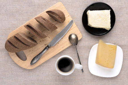 Sliced bread and knife on a cutting board. Butter, cheese, a cup of coffee on a sackcloth. Flat lay breakfast brunch on table conceptの写真素材