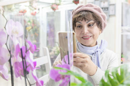 Happy caucasian elderly woman in a beret, smiling, photographs orchid flowers on the phone
