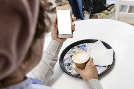 Caucasian woman in a beret, sitting at a table in a cafe, looking at the phone screen. In the...