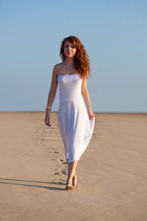 red-haired beautiful woman walking on sand wearing white dressの写真素材