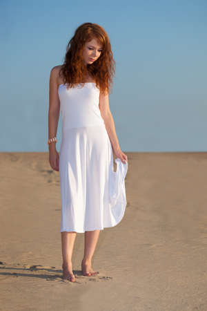red-haired beautiful woman walking on sand wearing white dressの写真素材