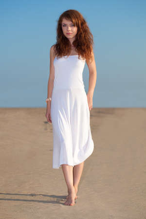 red-haired beautiful woman walking on sand wearing white dressの写真素材