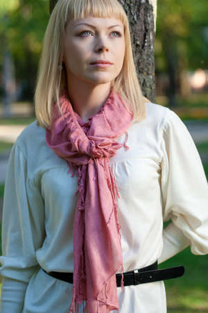 dreaming blond woman wearing pink scarf near tree in the summer parkの写真素材