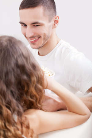 young couple sitting on the sofa eating popcorn and looking to each other, man laughの写真素材