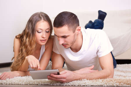 young couple lying on carpet and looking at mini tablet, woman pointing on displayの写真素材